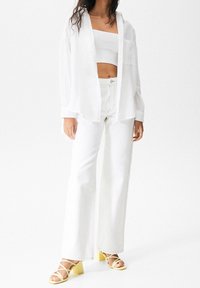 White linen button-up shirt layered over a white cropped top, paired with high-waisted white wide-leg jeans and yellow block-heeled sandals.