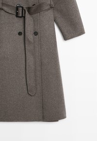 Cappotto di lana grigio con cintura in vita, chiusura doppiopetto con bottoni e maniche lunghe. Presenta una texture liscia e un design strutturato.