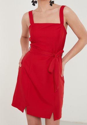 Kobieta w czerwonej sukience bez rękawów z kwadratowym dekoltem i związanym paskiem w talii, z rękami w kieszeniach, nosząca czarne kolczyki w kształcie kwiatów.