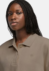 Beige polo tričko s výrazným límečkem a dvěma knoflíky, vyrobené z měkké látky s vyšitým logem na levém prsu.