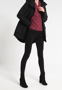 Black parka with a hood, maroon V-neck shirt, black skinny pants, and ankle boots. Features zippers and pockets on the parka.