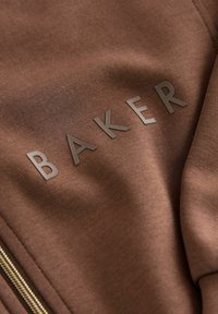 Bruine stof met het woord "BAKER" in een glanzende afwerking. Glad textuur met een ritsdetail dat zichtbaar is langs de rand.