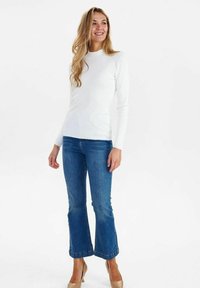 Weißes, geripptes Langarmoberteil mit hohem Ausschnitt, kombiniert mit blauen Schlagjeans und beigen High Heels; glattes Material und körperbetonter Schnitt.