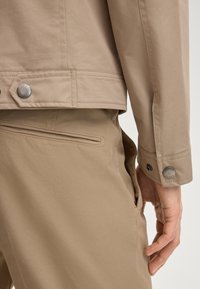 Zbliżenie osoby noszącej beżowe spodnie i dopasowaną kurtkę, ukazujące tylnią kieszeń, mankiet z markowym guzikiem oraz zrelaksowaną dłoń u boku.