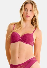 Lila kant beha met beugels en verstelbare schouderbandjes, met bloemmotieven en een kleine metalen accent in het midden. Gladde textuur.