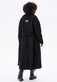 Czarny długi płaszcz wykonany z teksturowanej tkaniny, z paskiem w talii, z metką z tyłu. Długość do kolan, lekko luźny krój.