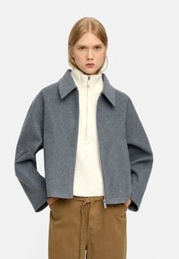 Szary żakiet z wełny z szerokim kołnierzem, zamek z przodu i obniżonymi rękawami, w połączeniu z jasnym swetrem na zamek i beżowymi spodniami.