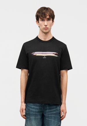 Homme portant un t-shirt noir avec un motif coloré de coups de pinceau horizontaux et un short en jean bleu, debout devant un fond clair uni.