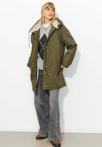 Parka vert olive avec une doublure en polaire beige confortable, une capuche à cordon de serrage, des poches avant et une coupe décontractée, assortie à un jean gris à jambes larges.