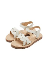 Sandales en cuir blanc avec un design de sangle tissée, des brides à la cheville réglables et une semelle plate brun clair. Aucun matériel supplémentaire visible.