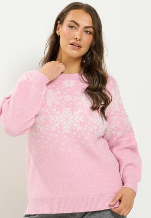 Femme aux longs cheveux ondulés portant un pull rose avec des motifs de flocons de neige blancs, regardant légèrement sur le côté et touchant son cou.