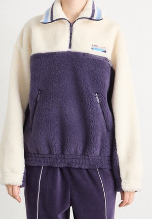 Person wearing a cream and navy blue fleece pullover with a zippered chest pocket and matching navy pants with white piping.