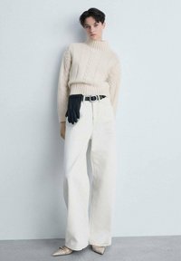 Beiger Kabelstrick-Turtleneck-Pullover, weiße weite Jeans, schwarzer Lederhandschuh am Bund und spitze beigefarbene Schuhe. Glatte Texturen.