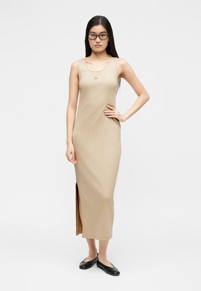 Γυναίκα με μακριά σκούρα μαλλιά και γυαλιά, φορώντας ένα μπεζ αμάνικο μακρύ φόρεμα με σκίσιμο στο πλάι και μαύρα επίπεδα παπούτσια, στέκεται μπροστά σε απλό φόντο.