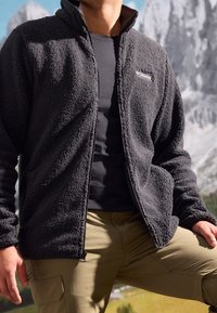 Giacca in pile nera con superficie texturizzata, con zip frontale e cappuccio, indossata sopra una maglietta scura e pantaloni cargo color khaki.