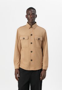 Veste beige en tissu doux, dotée d'un col, de deux poches avant à rabat et de boutons noirs, portée sur un pantalon sombre.