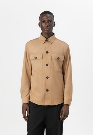 Tan jacket made of soft fabric, featuring a collared neck, two front pockets with flaps, and black buttons, worn over dark pants.