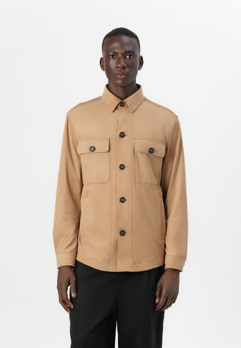 Veste beige en tissu doux, dotée d'un col, de deux poches avant à rabat et de boutons noirs, portée sur un pantalon sombre.
