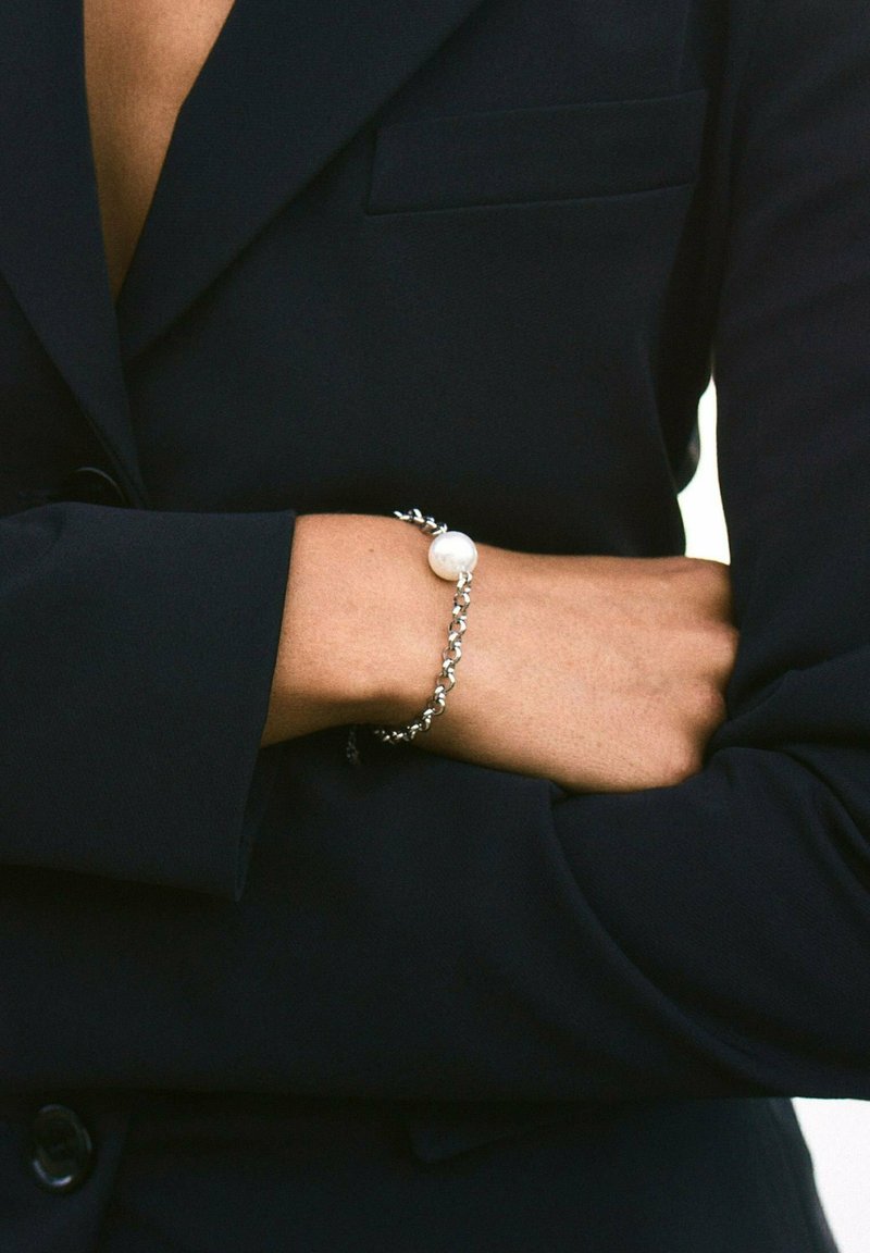 Brazo cruzado sobre el torso, vistiendo un blazer oscuro con una pulsera de cadena plateada que presenta una gran perla blanca.