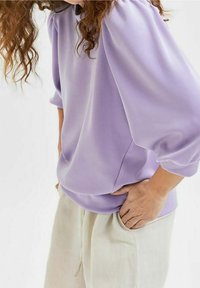 Jersey de color lavanda con mangas fruncidas, tejido suave, corte holgado, llevado sobre pantalones de color claro con bolsillos.