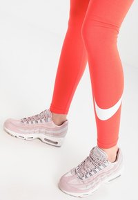 Osoba oblečená v jasně červených legínách s bílým logem Nike a světle růžových teniskách Nike Air Max stojí na bílé podlaze.