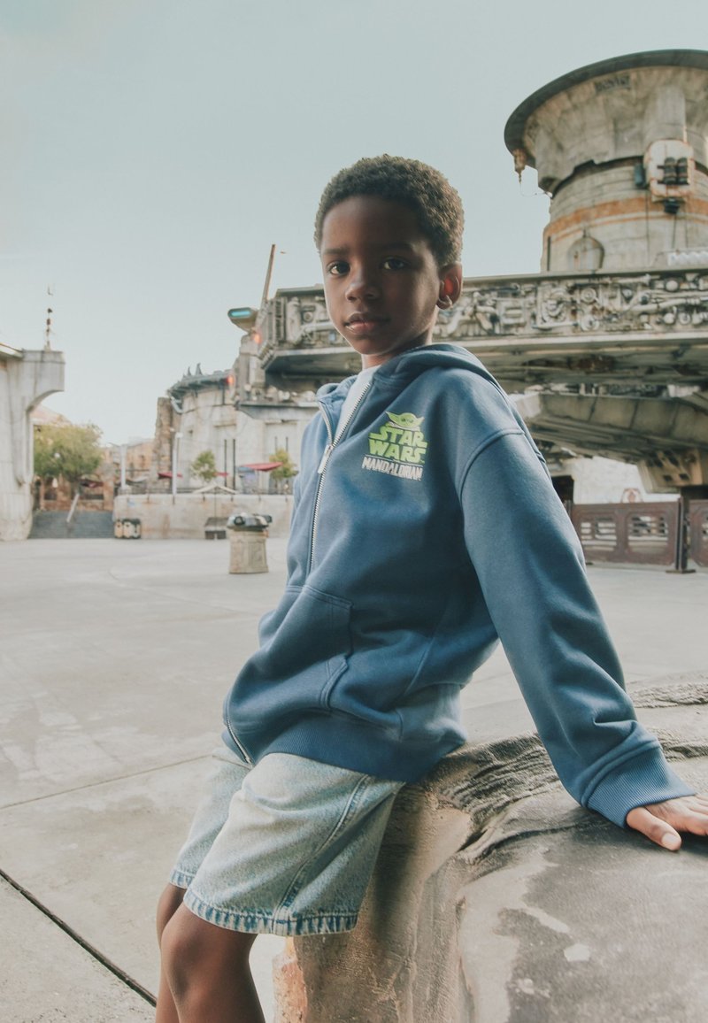 Young boy wearing a blue Star Wars Mandalorian hoodie and denim shorts sits on a stone ledge in a sci-fi themed outdoor area.