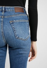 Jeans de denim azul con una forma ajustada, con un parche de cuero marrón en la cinturilla y un único bolsillo trasero con costuras amarillas.