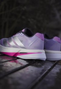 Fioletowe buty do biegania z srebrnymi akcentami, teksturowany górny materiał z siateczki oraz różową podeszwą. W designie znajdują się trzy paski i amortyzujący obcas.