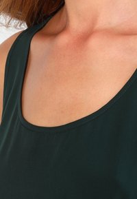 Haut vert foncé sans manches avec un col arrondi, un tissu lisse et des coutures soignées le long des bords, laissant la peau visible en dessous.