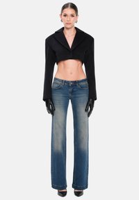 Chaqueta corta de lana negra con solapas anchas, combinada con vaqueros de cintura alta. La modelo lleva guantes negros y tacones de punta fina.