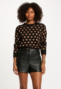 Maglione nero con motivi a cuore color rame, colletto rotondo e maniche lunghe, abbinato a pantaloni corti in pelle nera a vita alta. Capelli ricci e sfondo neutro.