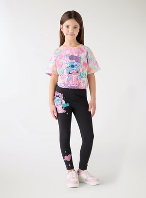 Młoda dziewczyna stoi w pastelowej koszulce tie-dye i czarnych legginsach z kolorowymi postaciami z kreskówek oraz napisem „You & Me.”