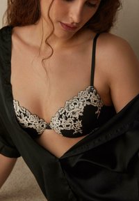 Svart spetsbehå med blommig broderi i krämfärgad nyans. Har en djup urringning och justerbara axelband. Kombineras med en svart satinblus som är delvis uppknäppt.