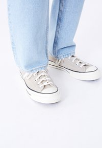 Světle šedé plátěné sneakers s bílými tkaničkami, gumovými podrážkami a černými akcenty, doplněné světle modrými džíny s rovnými nohavicemi.