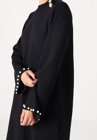 Person som har på sig ett svart långärmat plagg med pärlknappar på axlarna och pärldekorationer längs de utsvängda ärmsluten.