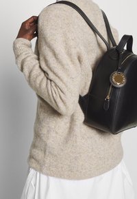 Mochila de cuero negro con forma redondeada, herrajes dorados y cierre de cremallera. Llevada a la espalda con un suéter de punto beige y una falda blanca.