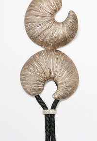Spilla in argento metallizzato con due forme spiraliche, decorate con linee testurizzate e un accento in pelle nera intrecciata, fissata con un fascetto in argento.