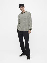 Jeune homme debout portant une chemise à manches longues rayée olive et blanche, pantalon noir et baskets noires avec semelles blanches sur fond blanc uni.