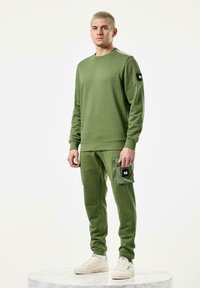 Olive green sweatshirt with long sleeves and ribbed cuffs, paired with matching joggers featuring zippered pockets and a textured patch.