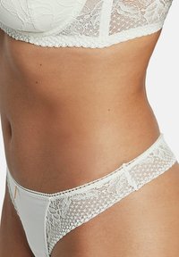 String en dentelle blanche avec une taille texturée. Présente des accents en dentelle florale et un corps en tissu lisse. Bords délicats en forme de fleur et quincaillerie dorée subtile.