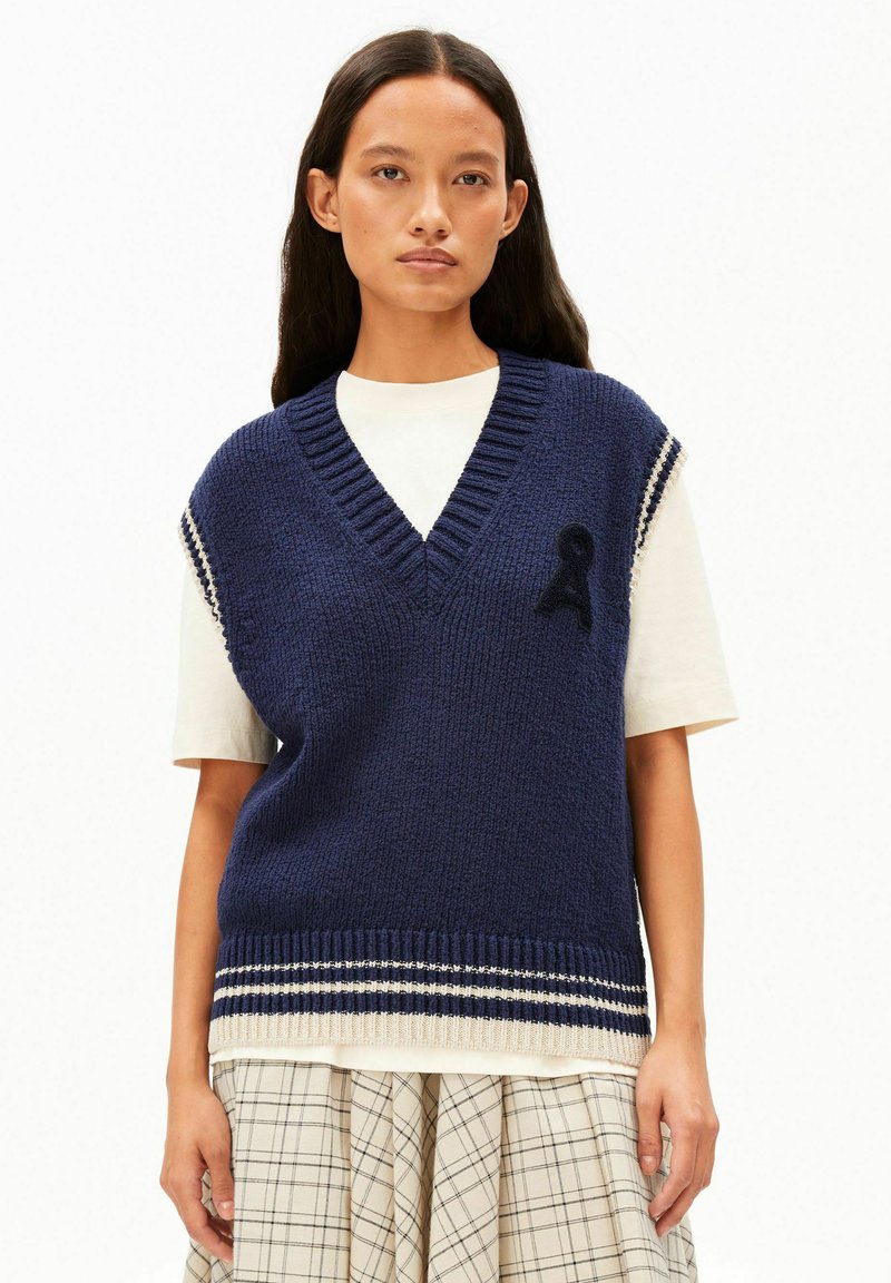 Gilet en tricot bleu marine avec un col en V profond, une bordure côtelée et des accents de rayures blanches. Présente une lettre noire brodée à l'avant.