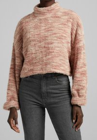 Różowy sweter crop z teksturą, z wysokim kołnierzem i długimi rękawami, noszony na ciemnoszarych dżinsach. Materiał ma miękkie, puszyste wykończenie.