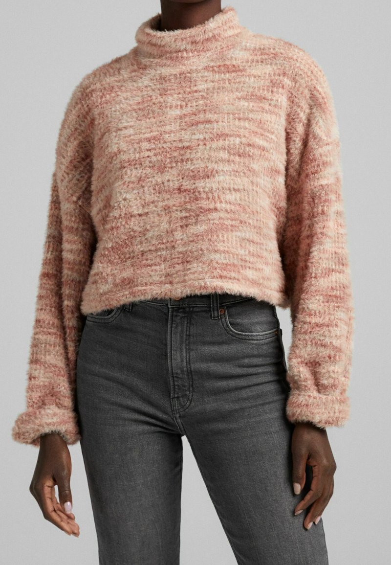 Różowy sweter crop z teksturą, z wysokim kołnierzem i długimi rękawami, noszony na ciemnoszarych dżinsach. Materiał ma miękkie, puszyste wykończenie.