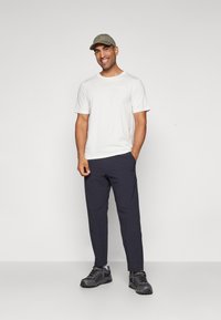 Weißes T-Shirt mit kurzen Ärmeln, marineblaue Hose, grüne Kappe und graue Wanderschuhe an einem Mann, der vor einem schlichten Hintergrund steht.