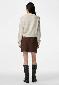 Woman facing away wearing a beige knit sweater, brown suede mini skirt, and black knee-high boots with buckles, standing on a white background.
