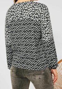 Der Pullover hat einen gemusterten Rücken in Schwarz und Weiß mit einer strukturierten Webart. Das Design umfasst eine Mischung aus abstrakten Formen und Zickzacklinien.