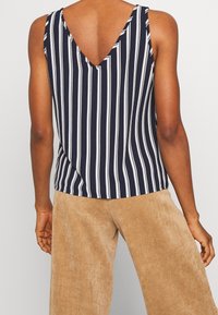 Personne portant un haut sans manches à rayures verticales bleu marine et blanches avec un profond dos en V, associé à un pantalon en velours côtelé beige.