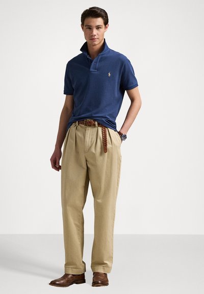 Marineblauwe polo met korte mouwen, kraag omhoog. Khaki geplooide broek met een gevlochten bruin leren riem. Brown dress shoes maken de look af.