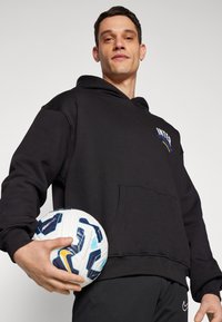Hoodie preto com um bolso frontal, apresentando o logótipo "INTER". Modelo segura uma bola de futebol branca e azul com padrão, vestindo calças de desporto pretas.