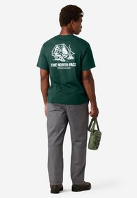 Camiseta de algodón verde con un gráfico blanco de una escena de camping en la parte trasera, combinada con pantalones grises y una pequeña bolsa verde oliva.